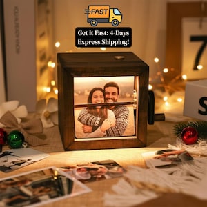May include: A dark wooden photo cube with a picture of a couple embracing inside. The cube has a crank on the side and is illuminated. The text "Get it Fast: 4-Days Express Shipping!" is displayed above the cube.