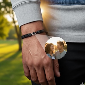 Könnte beinhalten: Schwarz geflochtenes Armband mit Silberverschluss und schwarzem Stein. Ein kleines rundes Bild eines Paares ist auf dem Bild überlagert. Das Armband wird am Handgelenk einer Person getragen.