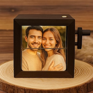May include: A dark brown wooden photo cube displaying a photograph of a smiling couple. The cube features a crank and rests on a wooden slice. The photo is visible through a rectangular aperture.