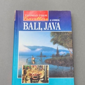 Può includere: Una guida di viaggio blu con il titolo "Thomas Cook Travellers Bali, Java & Lombok" in testo bianco. La copertina presenta una foto di un tempio tradizionale balinese su un lago con montagne sullo sfondo. C'è una foto più piccola di una maschera balinese nell'angolo in basso a sinistra. Il libro è etichettato "Thomas Cook" e "AA" nell'angolo in basso a destra.