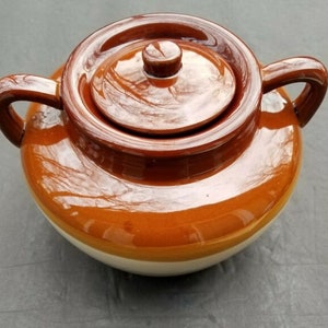May include: A brown glazed ceramic bean pot with a lid and two handles. The pot has a white band around the bottom.