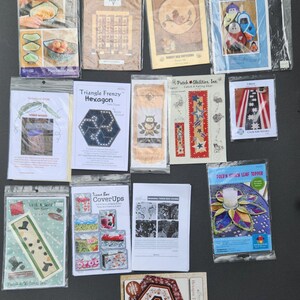 May include: A collection of 12 quilting patterns featuring various designs, including a welcome home sign, a triangle frenzy hexagon, a tissue box cover, a leaf topper, and a table topper. The patterns are printed on white paper and are packaged in clear plastic sleeves.