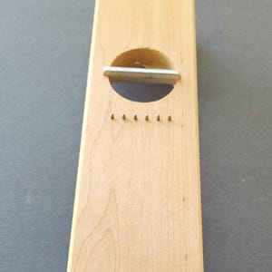 May include: A rectangular light-colored wooden kitchen tool featuring a circular cutout with a horizontal metal blade positioned across the opening, and a row of six small metal pins embedded in the wood surface below the blade.