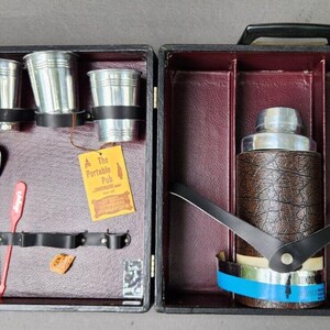 May include: A black leather carrying case with a silver cocktail shaker and six silver shot glasses. The case has a brown interior and a label that reads "The Portable Pub".
