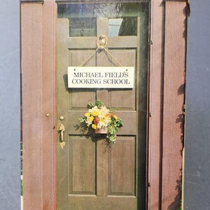 Puede incluir: Una portada de libro que presenta una puerta de madera con un letrero que dice "MICHAEL FIELD'S COOKING SCHOOL". Un pequeño arreglo floral cuelga debajo del letrero. La puerta está enmarcada por paneles verticales marrones.