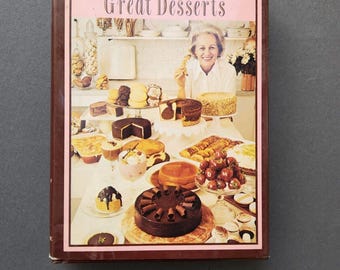 May include: A vintage cookbook titled "Maida Heatter's Book of Great Desserts" with a photo of a woman holding a dessert on the cover. The cover is pink with brown lettering and a border. The photo shows a table full of desserts, including cakes, pies, and pastries.