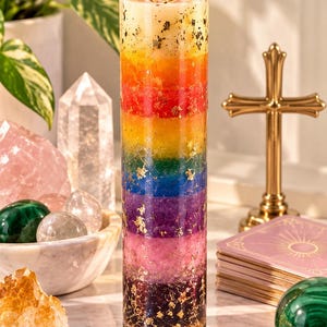 Puede incluir: Una vela alta y cilíndrica de vidrio con capas horizontales de cera en un espectro de colores arcoíris, incluyendo rojo, naranja, amarillo, verde, azul, morado y rosa, con copos de pan de oro incrustados en la cera y una mecha encendida.
