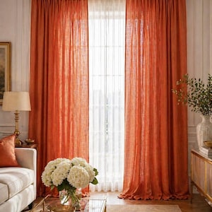 May include: Rust-coloured linen curtains hang from a gold rod, framing a window with white sheer panels. The room features a neutral colour scheme, with a sofa, coffee table, and decorative accents.