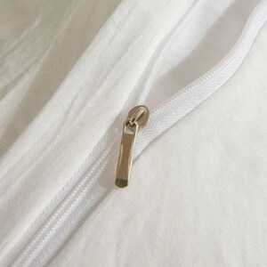 May include: Close-up of a white fabric with a zipper. The zipper is open, revealing the fabric's texture. The zipper pull is a gold color.