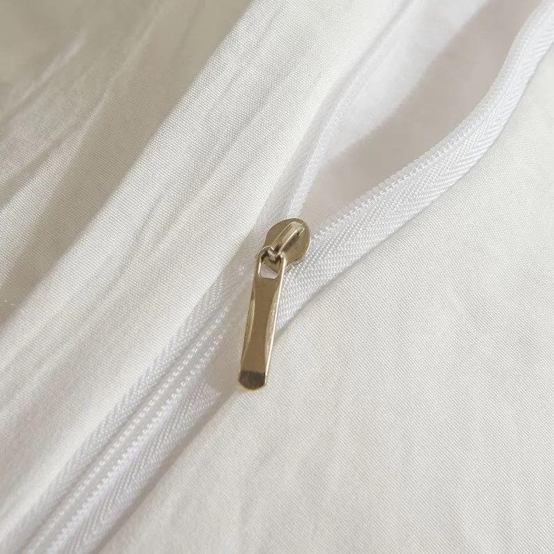 May include: Close-up of a white fabric with a zipper. The zipper is open, revealing the fabric's texture. The zipper pull is a gold color.