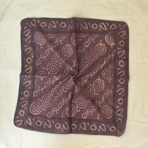 May include: A square, folded bandana in a deep purple hue. The fabric features a repeating pattern of cream-colored spirals and stylized floral designs. The edges are bordered with a decorative trim.