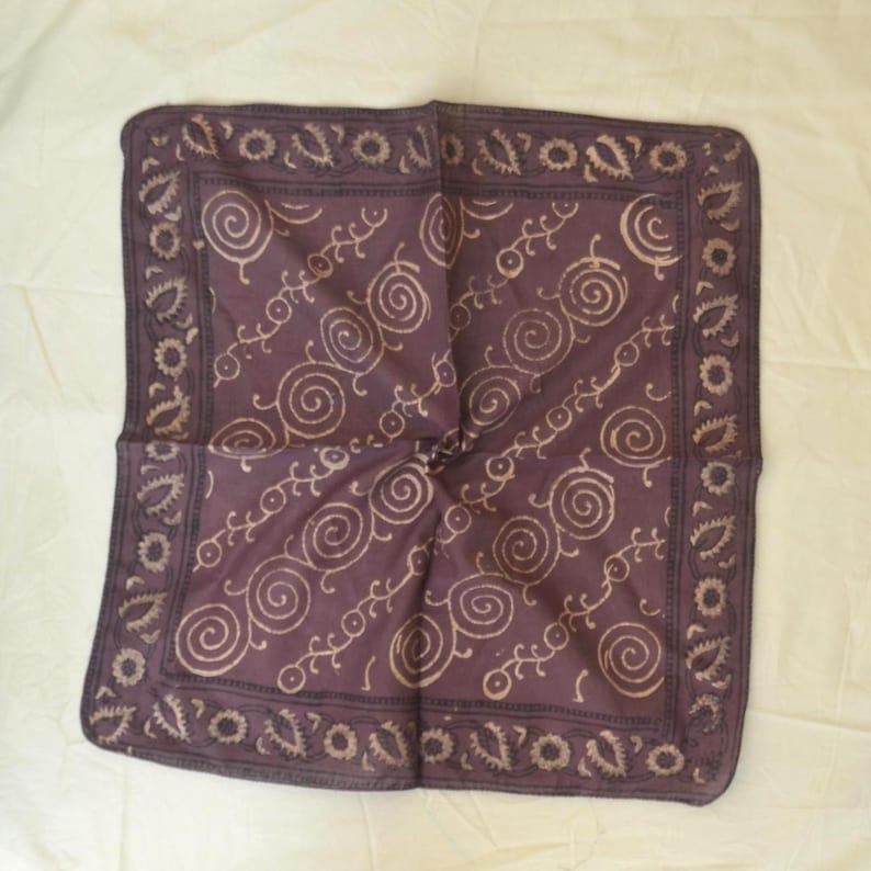 May include: A square, folded bandana in a deep purple hue. The fabric features a repeating pattern of cream-colored spirals and stylized floral designs. The edges are bordered with a decorative trim.