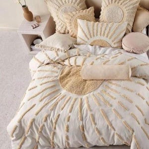 Puede incluir: Juego de cama color crema con diseño de sol. El juego incluye una funda nórdica, varias almohadas y un cojín cilíndrico. El diseño de sol se crea con detalles beige texturizados, agregando interés visual a la paleta de colores suaves y neutros.