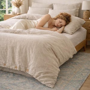 May include: A bed with beige linen bedding, including a duvet cover with wooden buttons. The bed is made up with pillows and a fitted sheet. A person is lying on the bed. The room has a neutral color palette with a rug.