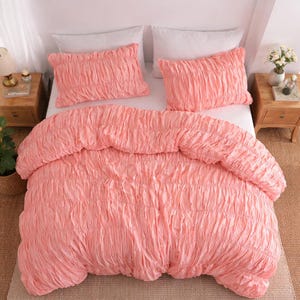 May include: A coral-colored bed set featuring a textured, ruched design. The set includes a comforter and two matching pillow shams. The bedding is arranged on a white bed, with wooden nightstands on either side.
