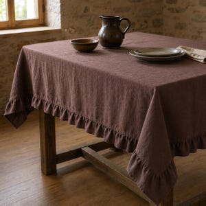 May include: A dark brown rectangular tablecloth with a ruffled edge draped over a wooden table. On the surface sits a dark brown ceramic pitcher, a small dark brown bowl, a stack of two ceramic plates, and a folded beige napkin.