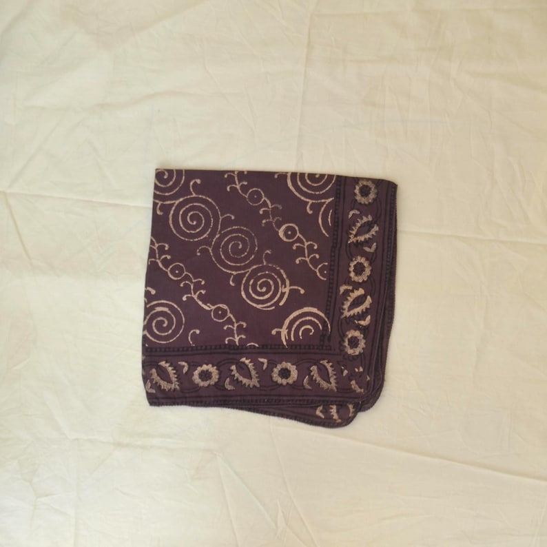 May include: A folded square scarf in a deep purple hue, adorned with a repeating pattern of cream-colored swirls and floral-like motifs. The edges feature a decorative border, creating a contrast against the solid background. The scarf is made of a soft fabric.