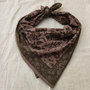 Könnte beinhalten: Ein dreieckiges Bandana mit einer staubig malvenfarbenen Grundfarbe und einem dunkelbraunen Blumenmuster. Der Stoff hat einen breiten, olivgrünen Rand mit einem detaillierten Blumendruck. Das Bandana ist oben mit einem Knoten gebunden.