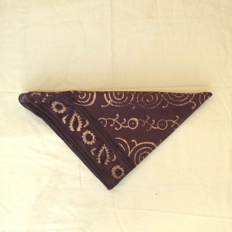 May include: A dark purple bandana with a beige floral and swirl pattern. The bandana is folded in a triangle shape, with the patterned border along one edge. The fabric appears to be cotton or a similar material.
