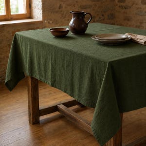 Puede incluir: Un mantel rectangular de color verde oliva oscuro con una textura tejida visible sobre una mesa de madera. En la superficie hay una jarra de cerámica marrón oscuro, un pequeño cuenco de madera a juego, dos platos de cerámica apilados y una servilleta beige doblada.