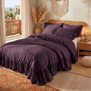 May include: A bed with a deep purple ruffled duvet cover and matching pillowcases. The bed is in a room with a woven headboard, a wooden nightstand, and a patterned rug. A macrame wall hanging and pampas grass add to the decor.