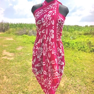 Op de afbeelding: Een felroze sarong met een wit bloemmotief is gedrapeerd op een mannequin. De sarong is om de torso van de mannequin gewikkeld en bij de nek vastgeknoopt. De achtergrond toont een groen veld en een bewolkte lucht.