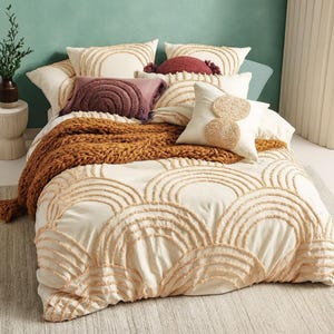 May include: A bed with a cream-colored comforter and pillows. The comforter features a repeating arch design in a lighter cream color. Several decorative pillows in shades of cream, burgundy, and brown are arranged on top, along with a chunky knit throw.