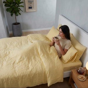 Könnte beinhalten: Ein Bett mit gelber Bettwäsche, einschließlich Bettbezug, Kissen und Spannbettlaken. Eine Person sitzt im Bett und hält eine Tasse. Ein kleiner Nachttisch aus Holz mit einer Lampe und einer Teekanne steht neben dem Bett.