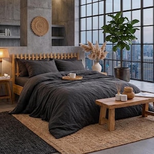 May include: A contemporary bedroom featuring a dark grey bedspread and pillows on a wooden bed frame. A small wooden table holds a tray with a cup and a decorative vase. A large window offers a city view.