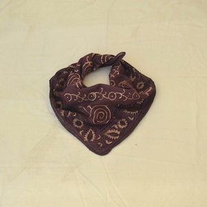 May include: A dark purple bandana with a paisley and floral pattern in a lighter tan color. The bandana is folded in a triangle shape, with the design visible on the front. The fabric appears to be soft and lightweight.