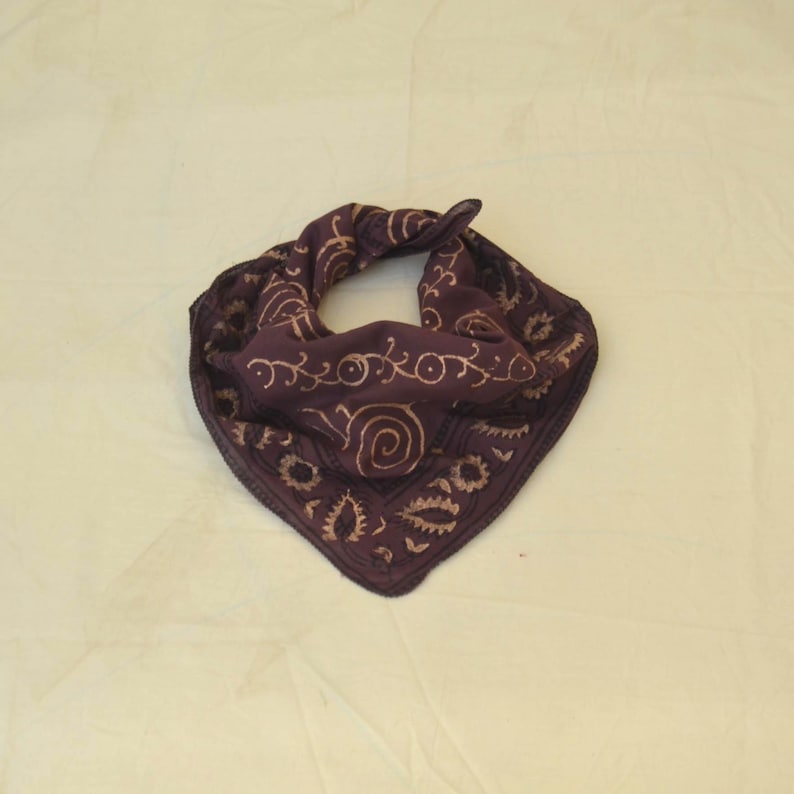 May include: A dark purple bandana with a paisley and floral pattern in a lighter tan color. The bandana is folded in a triangle shape, with the design visible on the front. The fabric appears to be soft and lightweight.