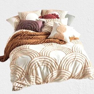May include: A bed with a white comforter featuring a repeating arched design in a light brown color. The bed is adorned with several pillows in shades of white, brown, and burgundy, and a chunky knit brown throw blanket.