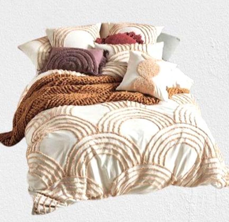 May include: A bed with a white comforter featuring a repeating arched design in a light brown color. The bed is adorned with several pillows in shades of white, brown, and burgundy, and a chunky knit brown throw blanket.