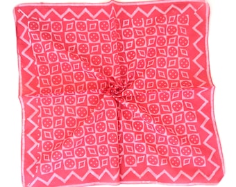 May include: A square, coral-coloured scarf with a repeating pattern of white circles, squares, and diamonds. The edges feature a white zigzag design. The scarf is folded in the centre.