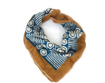 May include: A folded scarf with a blue and white patterned design on a tan background. The pattern features circles and lines. The scarf is made of a soft material and is displayed on a white background.