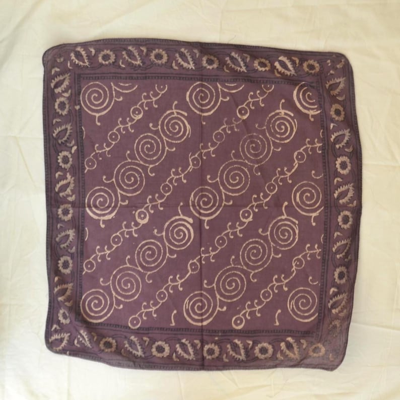 May include: A square, purple bandana with a repeating spiral and vine pattern in a lighter shade of purple. The border features a paisley and floral design. The fabric is soft and appears to be cotton.
