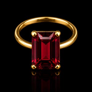 Pode incluir: Um anel de ouro com uma grande pedra preciosa vermelho-escura de corte retangular esmeralda, montada em quatro garras. A banda é lisa e polida, exibindo uma pedra carmesim vibrante que reflete a luz contra um fundo preto sólido.