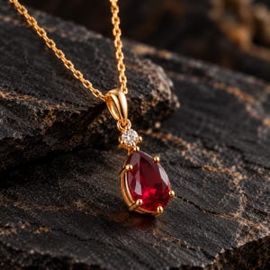 May include: A gold necklace featuring a pear-shaped red gemstone pendant suspended from a delicate chain. A small, round, clear gemstone is set directly above the red pear-shaped stone, held in place by a gold prong setting.