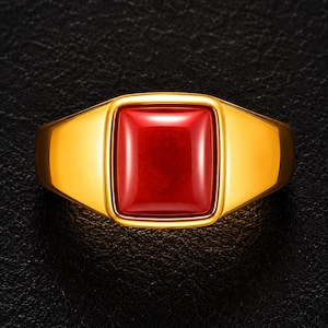 May include: A gold ring featuring a smooth, rectangular, deep red gemstone set in the center. The band is polished and solid gold, with a wide, tapered design that frames the vibrant red stone, creating a bold and clean aesthetic for the piece.