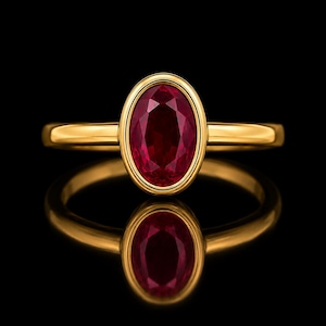 Puede incluir: Un anillo de oro con un rubí rojo intenso de talla ovalada engastado en un bisel liso. La banda es de oro amarillo pulido con un diseño sencillo y limpio, que resalta la vibrante piedra roja sobre una superficie oscura y reflectante.