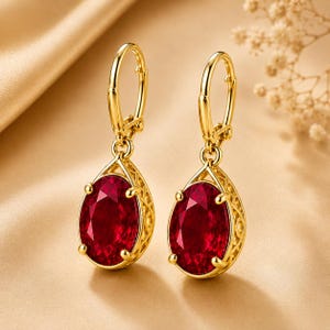 May include: A pair of gold leverback earrings featuring oval-cut deep red gemstones. Each stone is held by four prongs within a decorative gold frame with an intricate lattice pattern, suspended from a polished gold hoop mechanism.