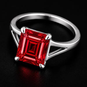 Puede incluir: Un anillo de tono plateado con una gran piedra preciosa roja vibrante de corte cuadrado, sujeta por un engaste de cuatro garras. La banda tiene un diseño de vástago dividido que se estrecha hacia atrás, creando un aspecto geométrico pulido.