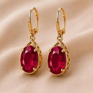 May include: A pair of gold leverback earrings featuring oval-cut, deep red faceted stones. Each stone is secured by four prongs within a decorative, openwork gold cage setting that tapers to a point at the top, connecting to a hinged gold loop.