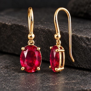 Puede incluir: Un par de pendientes colgantes de oro con piedras preciosas de color rojo vibrante de corte ovalado. Cada pendiente cuelga de un gancho de oro curvado y está sujeto con cuatro garras, mostrando un acabado metálico pulido y facetado.