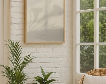 May include: A framed artwork with a light wood frame hangs on a white brick wall. The artwork features a soft, blurred image of flowers and foliage. Two potted plants and a wicker chair with a cushion are in the foreground.