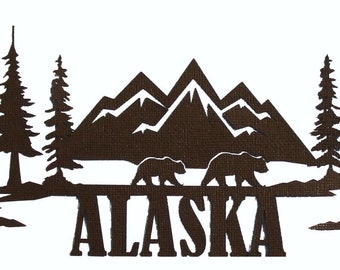 Może przedstawiać: Brązowy wycięty wzór z napisem "ALASKA" na dole. Nad tekstem znajdują się dwa niedźwiedzie, góry i drzewa. Projekt jest na białym tle.