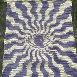 Puede incluir: Un tapiz de pared hecho a mano con un diseño de ilusión óptica. La pieza utiliza hilo lavanda y blanquecino para crear un patrón central en forma de ojo con rayas onduladas radiantes. Se utiliza una clavija de madera y una cuerda para colgar.