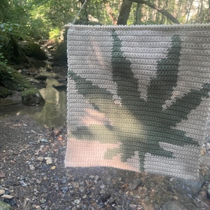 Peut inclure: Une tenture murale au crochet faite à la main, représentant une grande feuille de cannabis. La feuille est réalisée dans des tons de vert et de beige, sur un fond neutre. La pièce est suspendue par une cheville en bois et une ficelle, idéale pour la décoration.