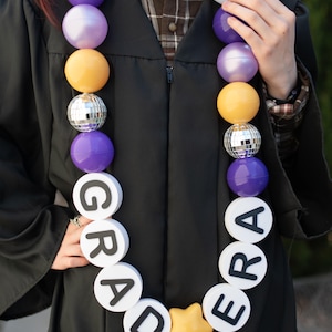 May include: A graduation lei featuring purple, light purple, and yellow spheres, silver disco ball beads, and white circular discs with black letters spelling GRAD ERA, accented by a yellow star charm at the bottom center of the necklace.