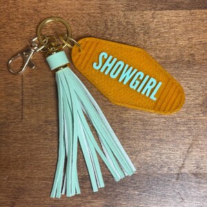 May include: A gold-colored motel-style keychain with the word "SHOWGIRL" in turquoise. The keychain has a gold ring, a mint green tassel, and a lobster clasp.